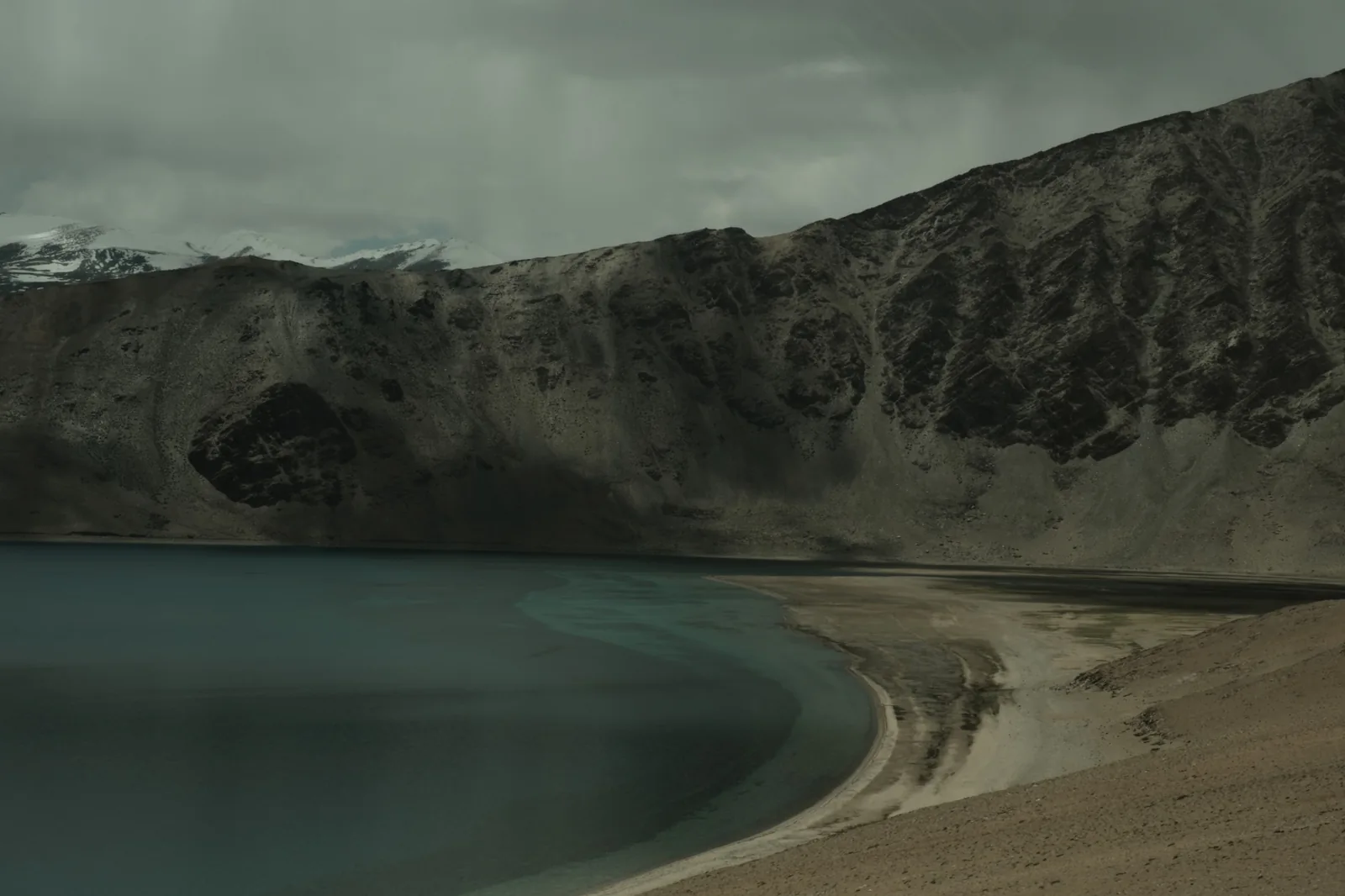 A dark lake curving along a sandy shore beneath steep, shadowed cliffs and grey cloud