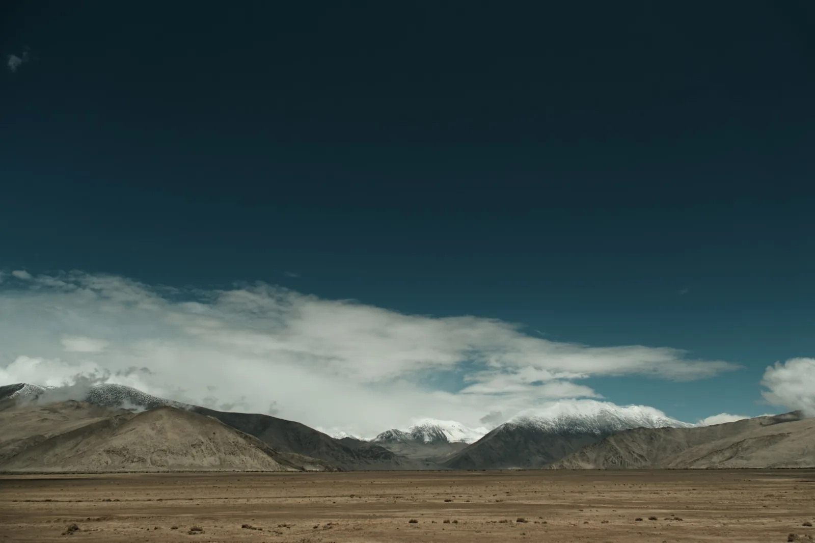 A wide, dry plain under a deep blue sky, snow-capped peaks and a long band of cloud on the horizon