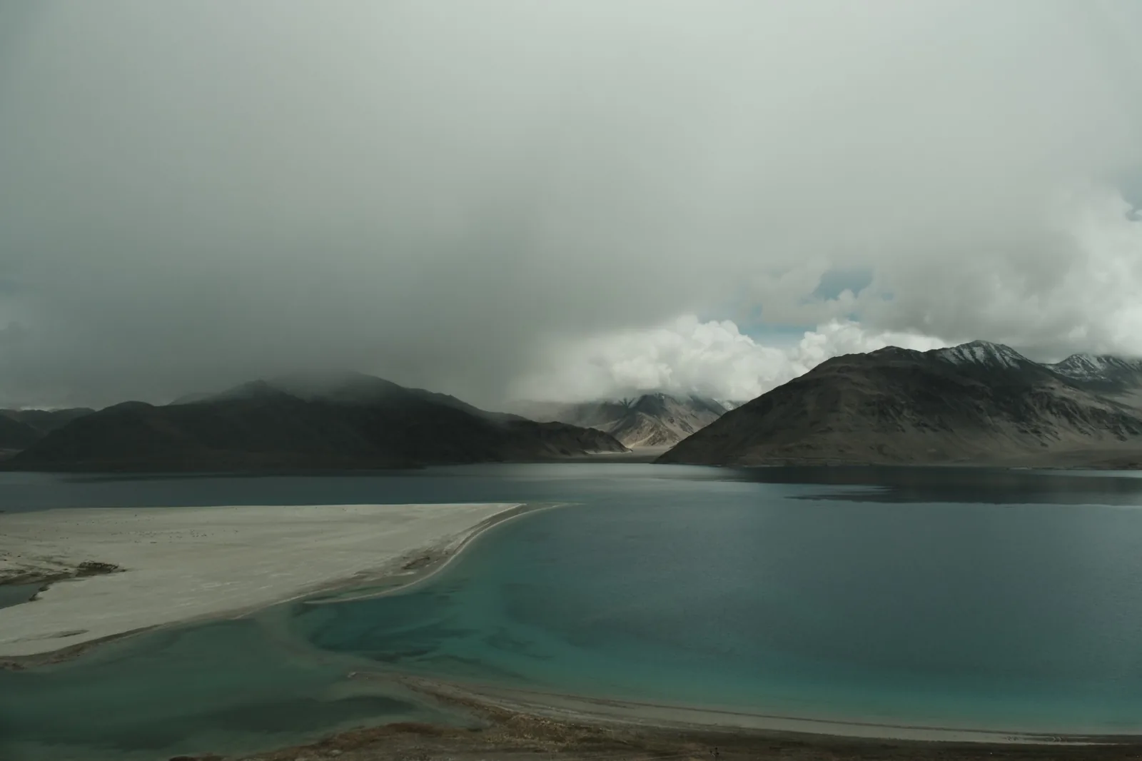 A turquoise lake under heavy cloud, a pale sandbar curving into it, dark mountains behind